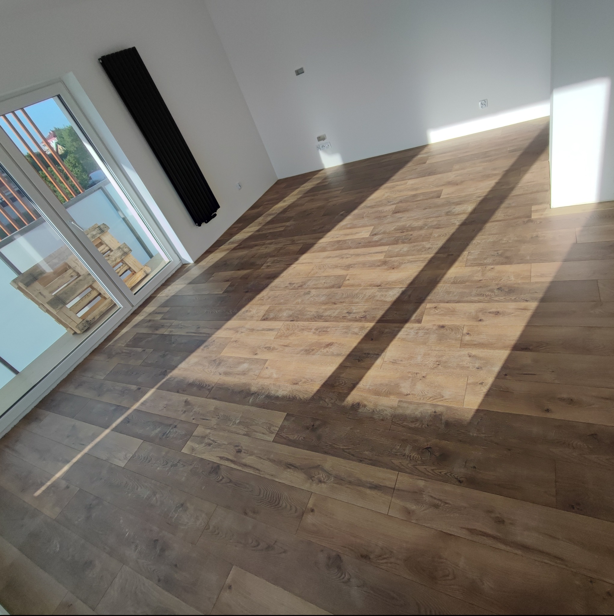 Ukończona podłoga z paneli w nowym mieszkaniu, widoczne promienie słońca wpadające przez okno balkonowe, przy oknie ustawione palety budowlane, na ścianie zamontowany czarny grzejnik.