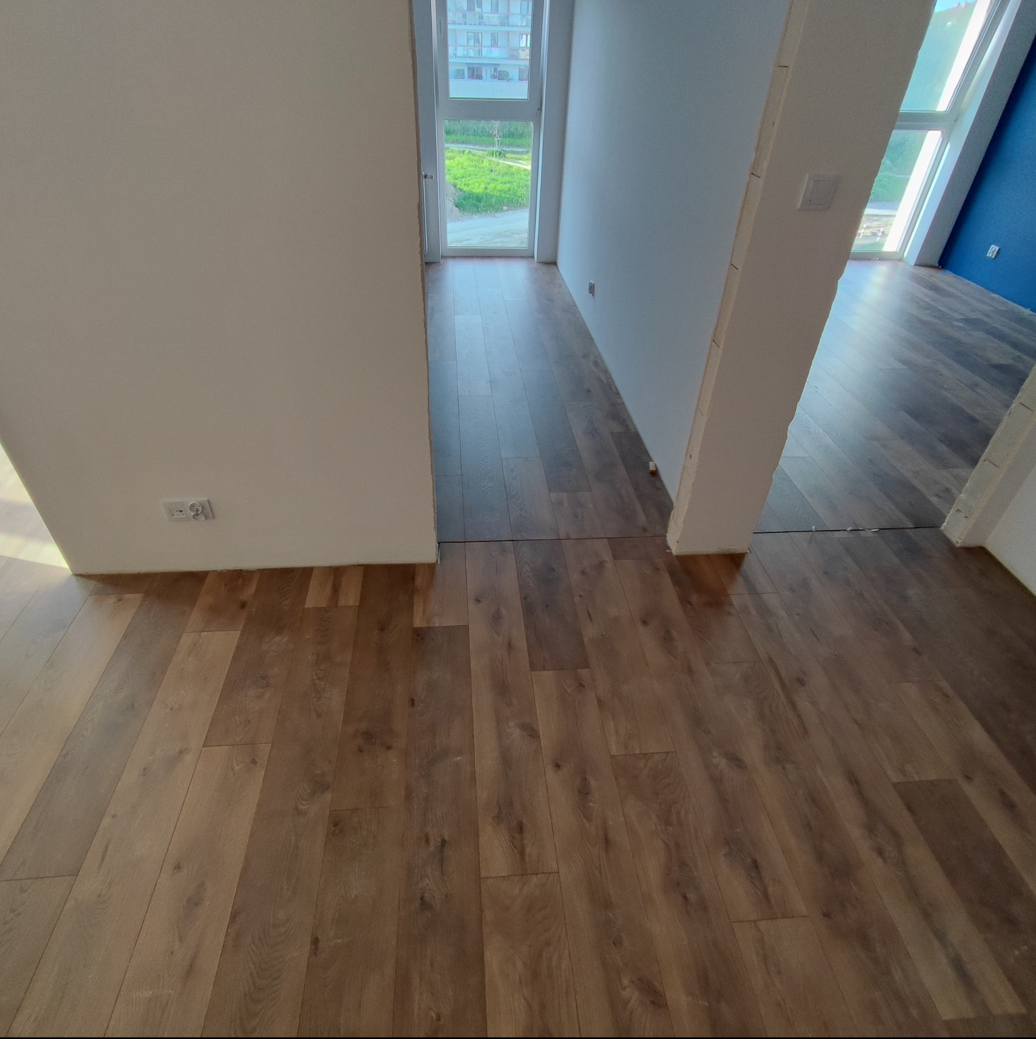 Ułożone panele podłogowe w nowym mieszkaniu, widoczne gniazdka elektryczne przy ścianie i fragment niebieskiej ściany w oddali.