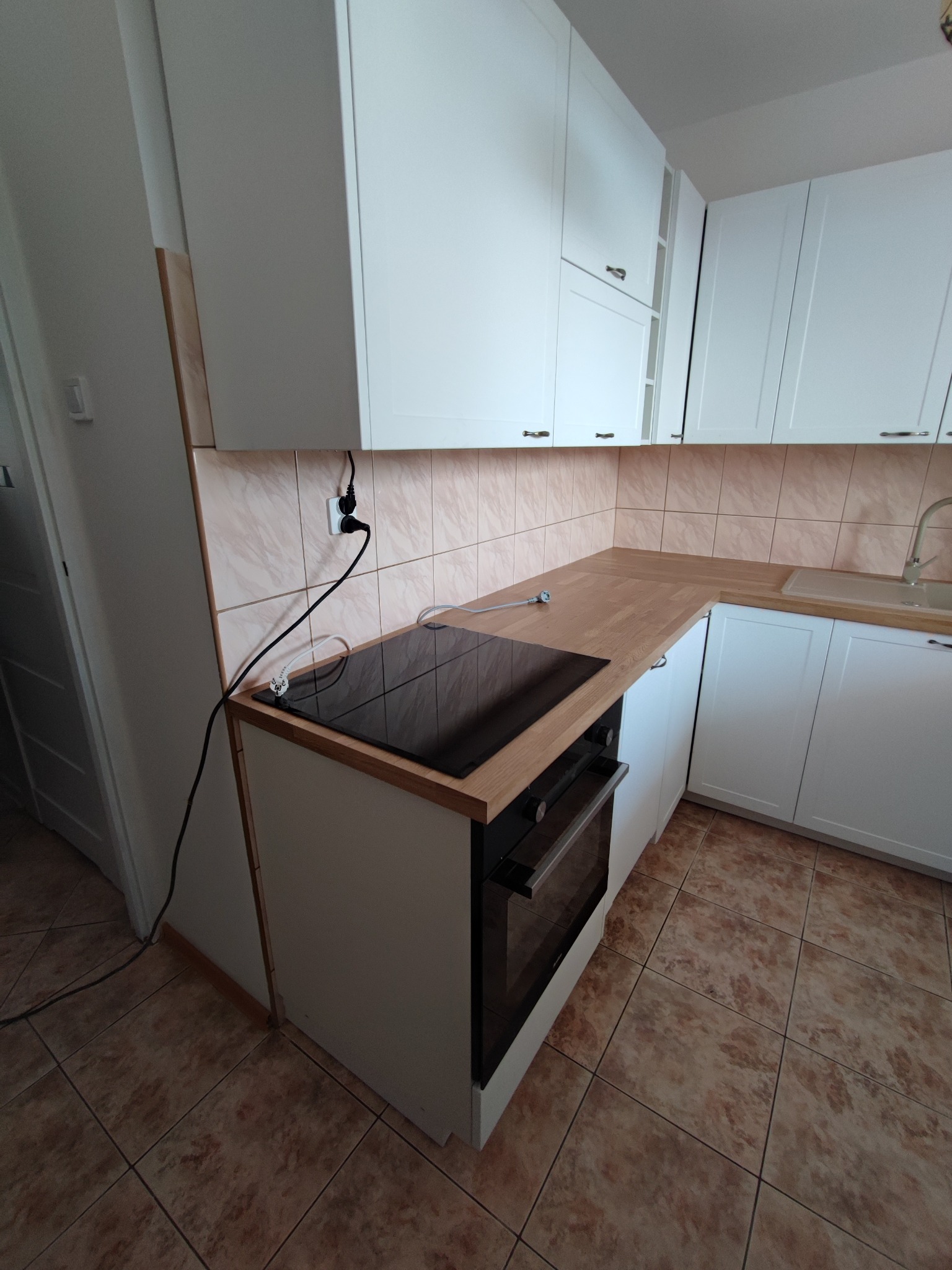 Nowoczesna kuchnia z białymi szafkami, drewnianym blatem i czarną płytą indukcyjną oraz piekarnikiem w zabudowie, widoczne gniazdko elektryczne i brązowe płytki podłogowe.