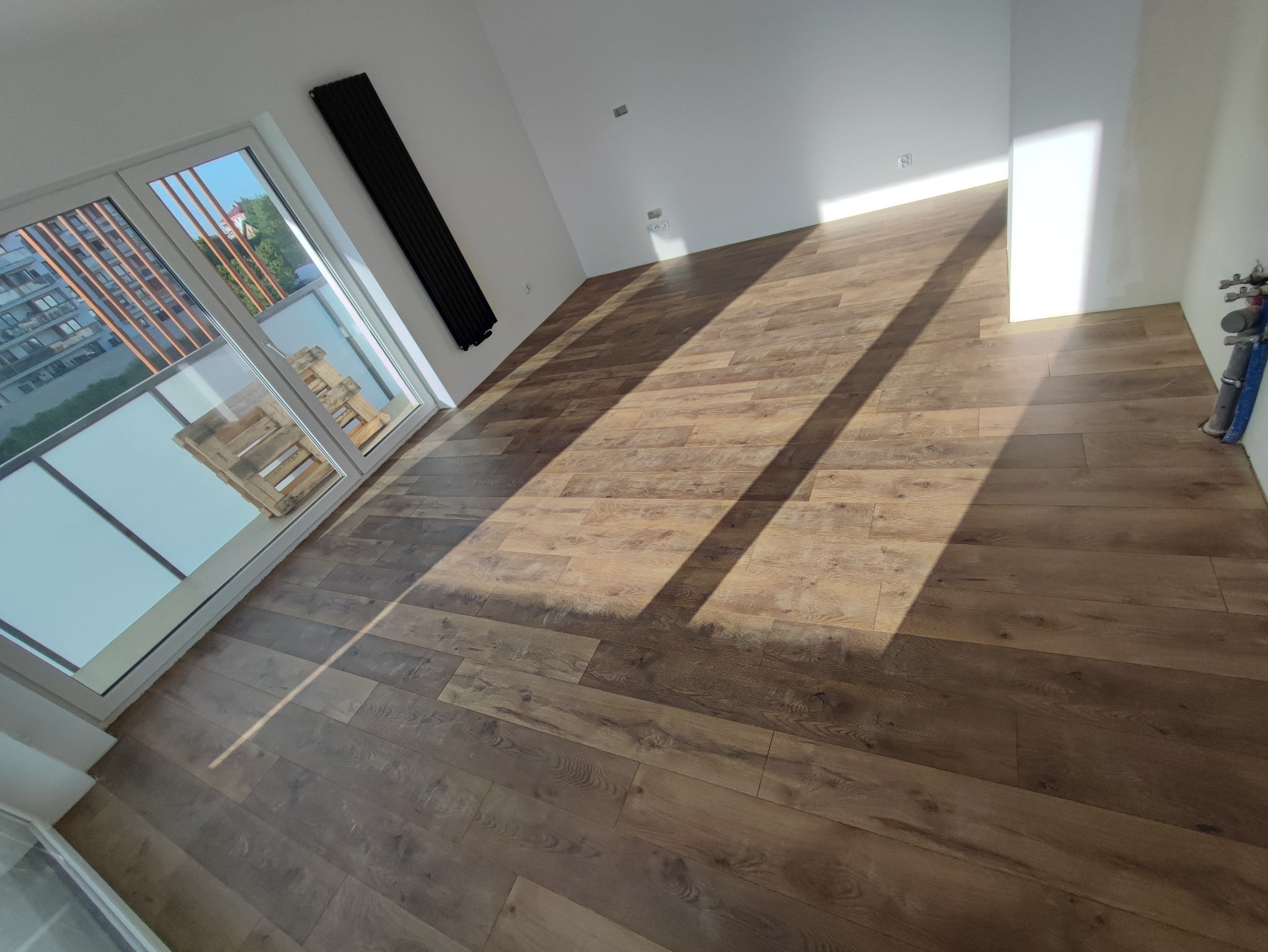 Nowo ułożona podłoga z paneli w jasnym pomieszczeniu, z widocznymi promieniami słońca, grzejnikiem na ścianie i paletą na balkonie za oknem.