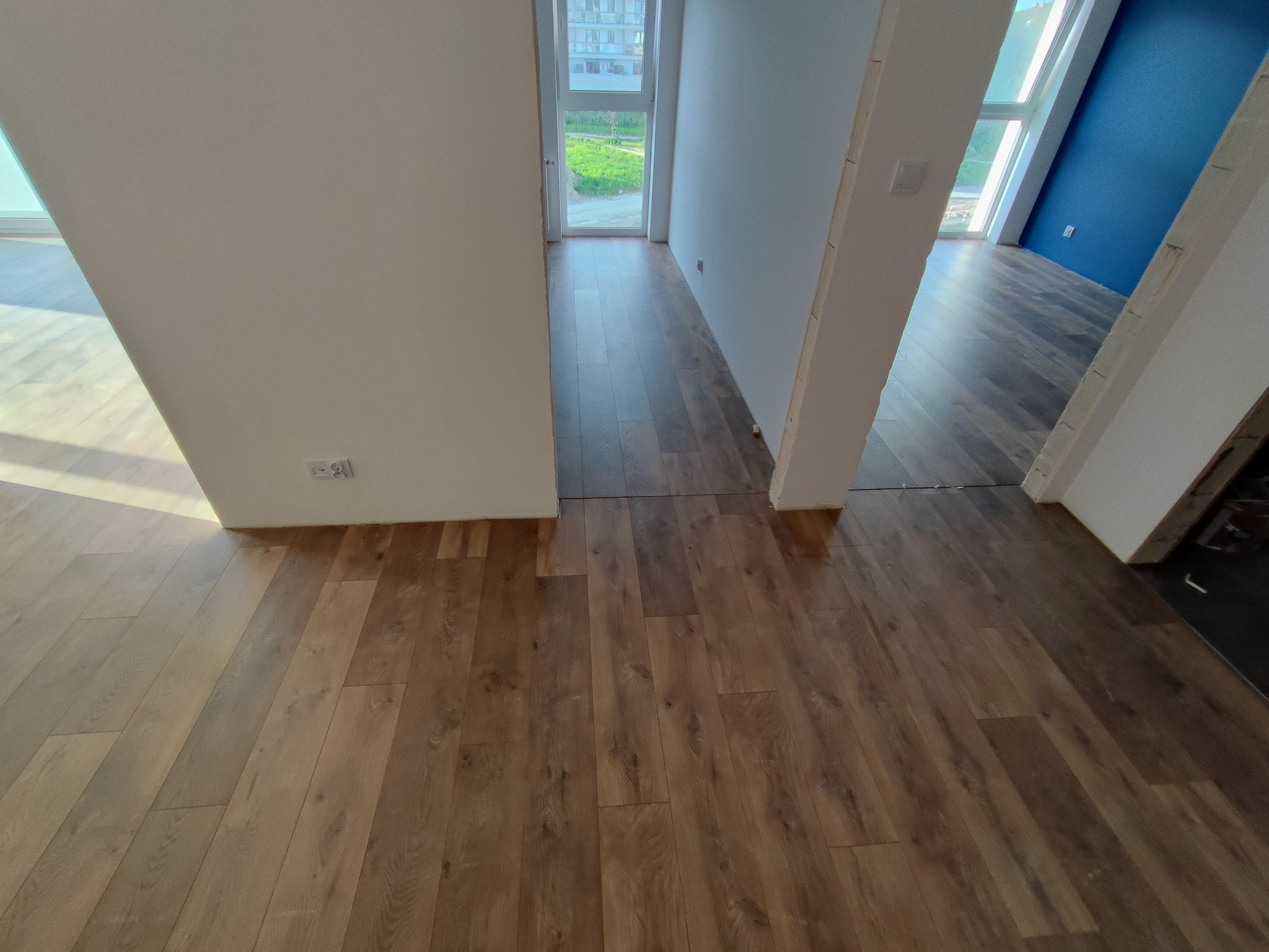 Ułożone panele podłogowe w odcieniach brązu w nowym mieszkaniu, widoczne przejścia między pomieszczeniami i fragment niebieskiej ściany w tle.