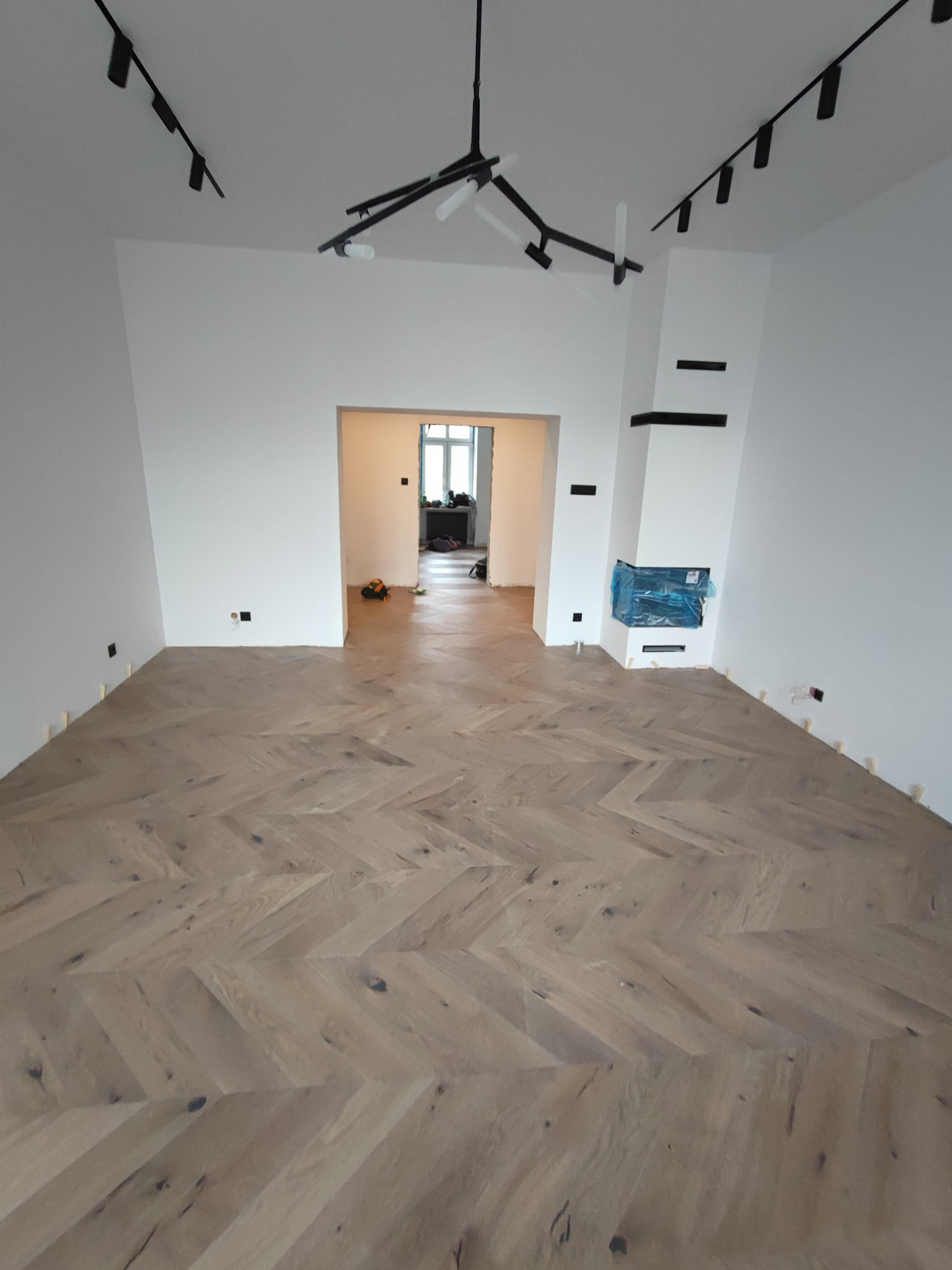 Jasny parkiet ułożony we wzór jodełki w przestronnym, białym wnętrzu z minimalistycznym oświetleniem i kominkiem zabezpieczonym folią.