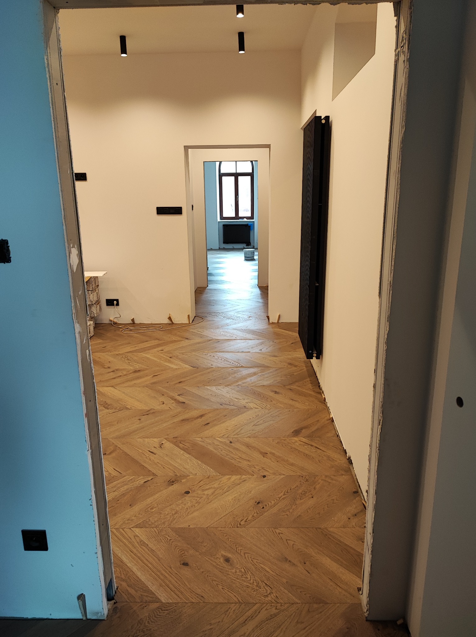 Wnętrze mieszkania w trakcie remontu, widok na ułożony parkiet w jodełkę, z widocznymi niedokończonymi pracami wykończeniowymi przy ścianach i ościeżnicach drzwiowych.