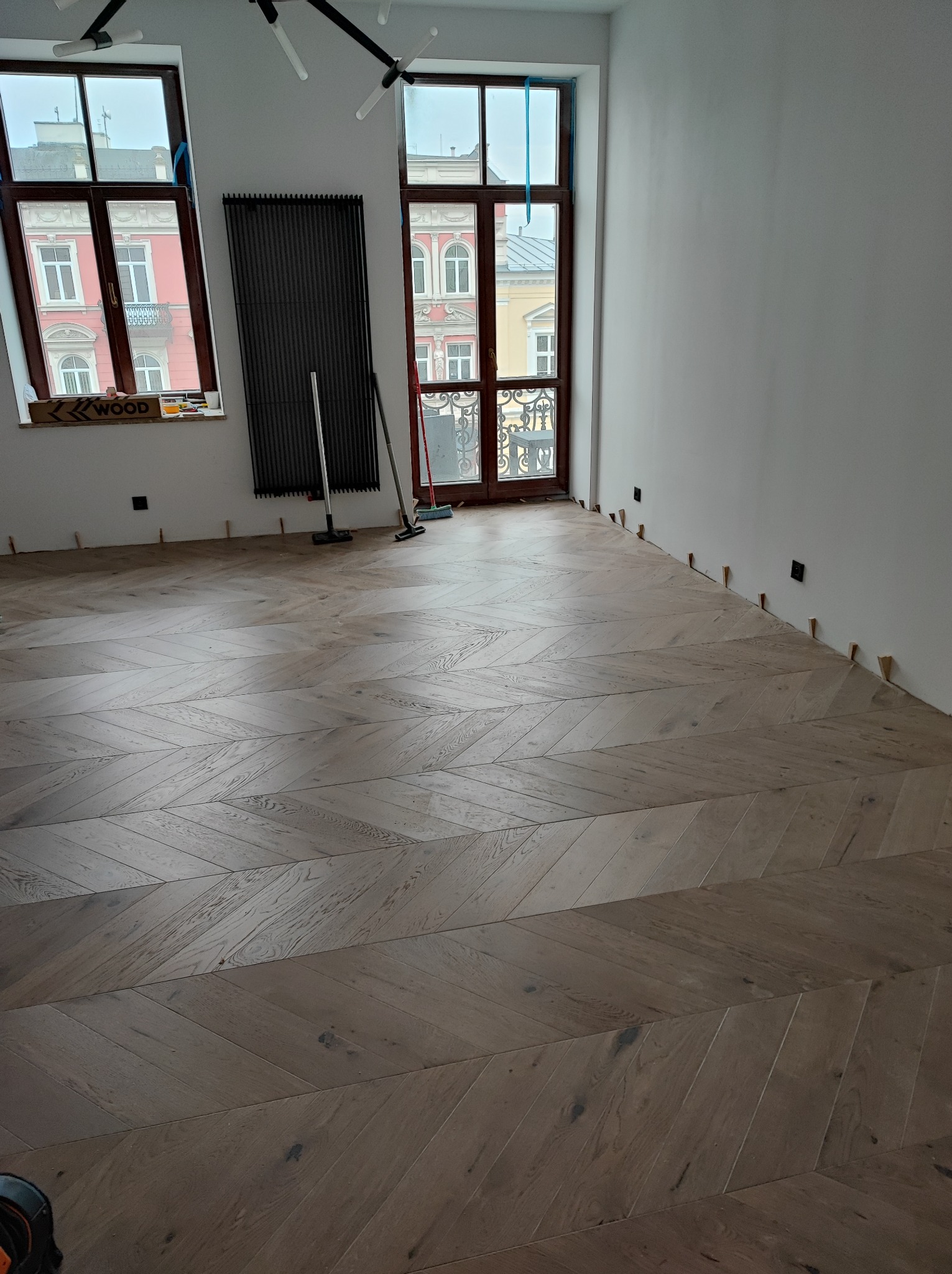 Ułożony parkiet w jodełkę w jasnym pomieszczeniu z dwoma oknami; widoczne kliny dystansowe przy ścianach i grzejnik dekoracyjny.