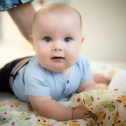 Niemowlę w błękitnym ubranku leży na brzuszku na kocyku w sowy, spoglądając w obiektyw z lekkim uśmiechem, w tle fragment kwiecistej bluzki osoby dorosłej.