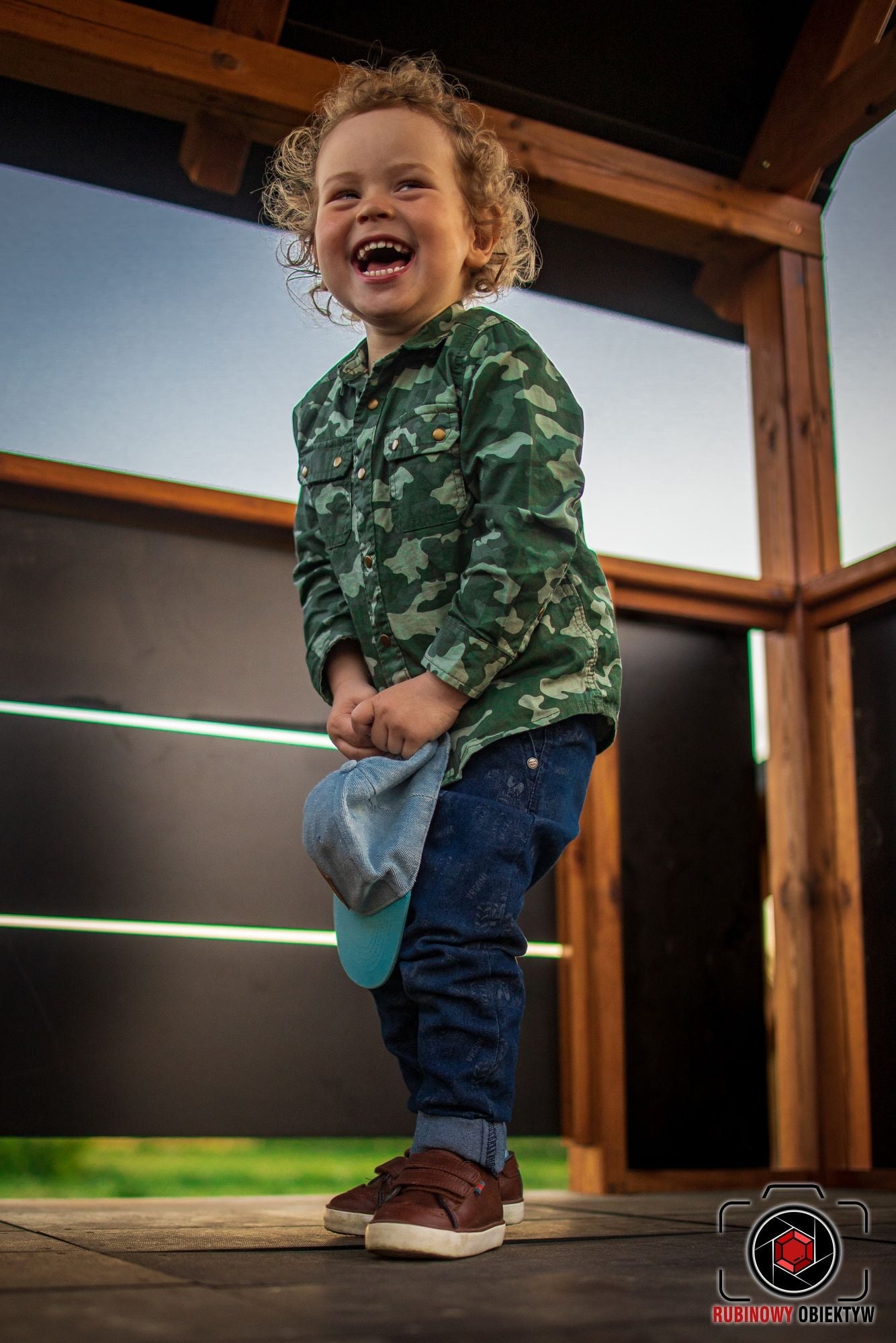 Uśmiechnięty chłopiec z kręconymi włosami w koszuli moro trzymający czapkę z daszkiem, stojący w drewnianej konstrukcji.