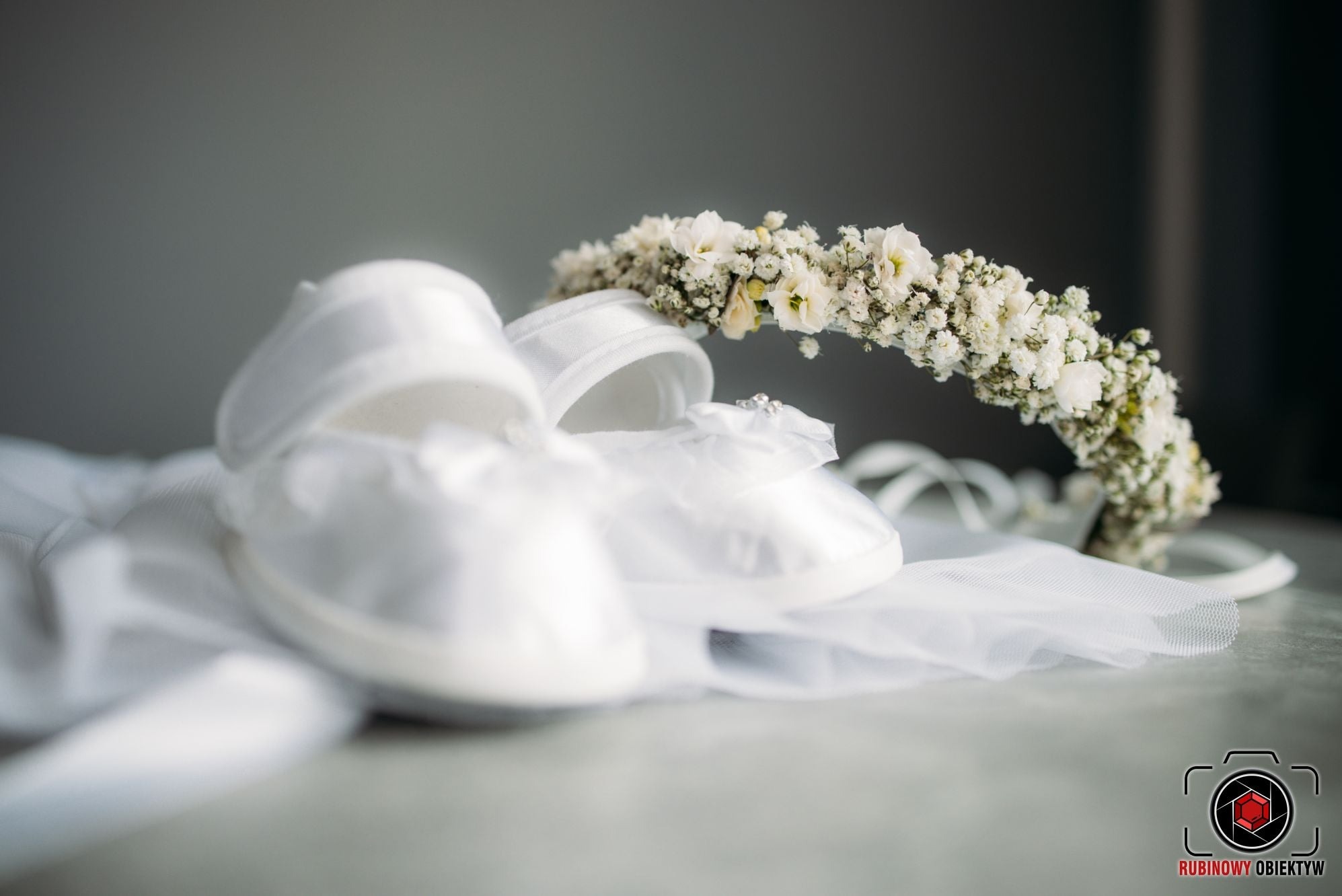 Białe satynowe buciki niemowlęce z kokardką i wianek z białych kwiatów, przygotowane do chrztu w Wysokiem Mazowieckiem, leżą na delikatnej siateczkowej tkaninie.