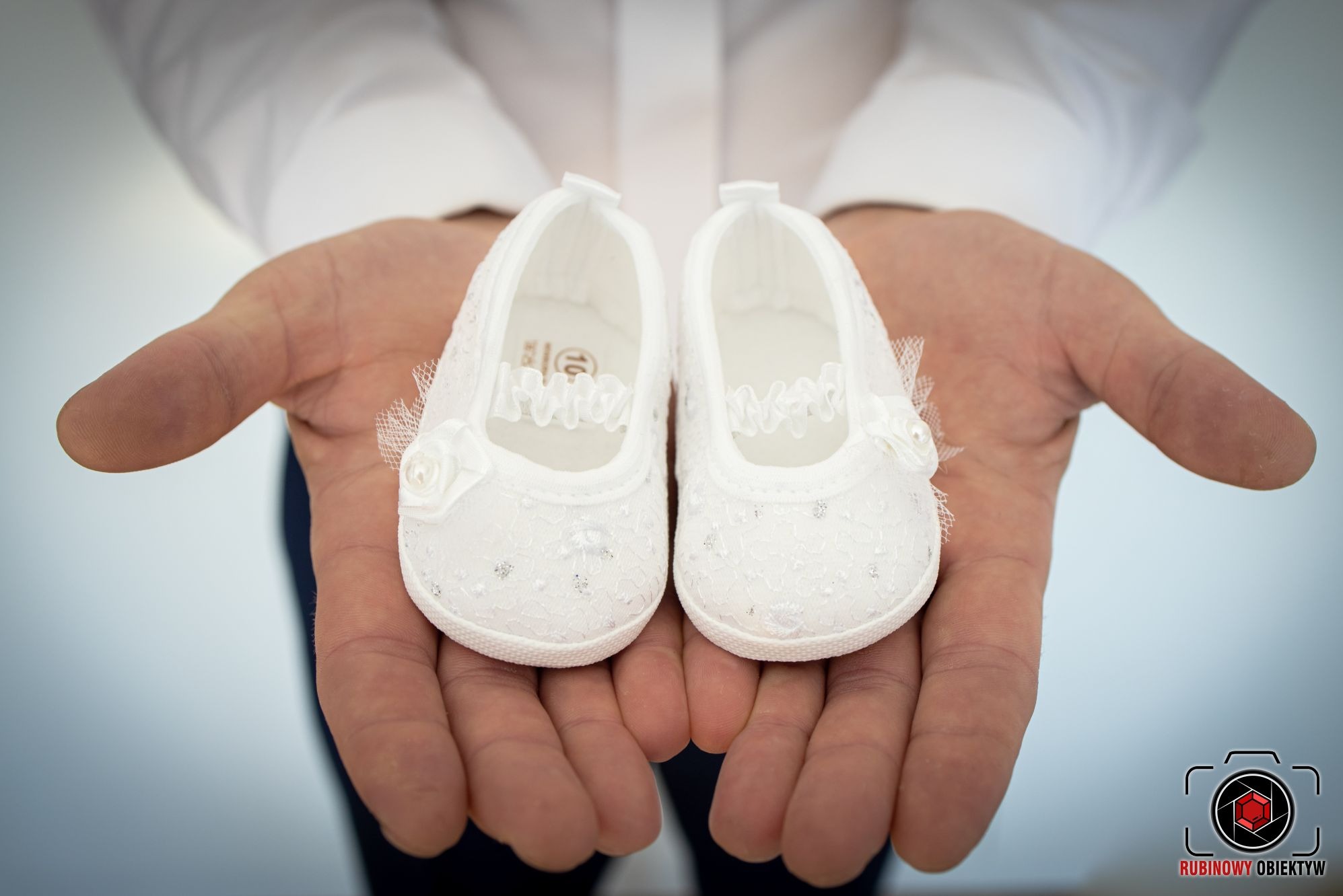Dłonie trzymające parę białych, koronkowych bucików niemowlęcych z ozdobnymi różyczkami, na jasnym tle.