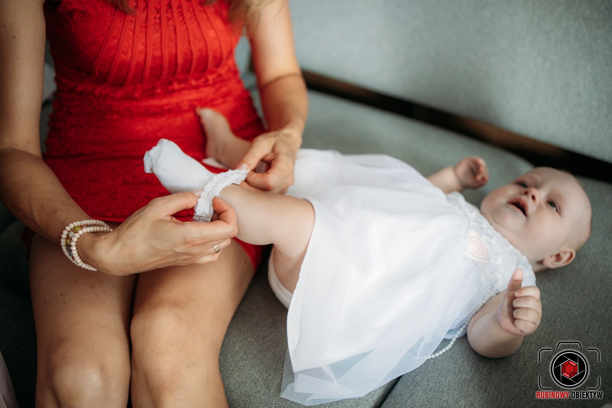 Mama w czerwonej sukience zakłada białą skarpetkę z koronką na nóżkę leżącego na szarym tle niemowlaka w białej sukience z perłami.