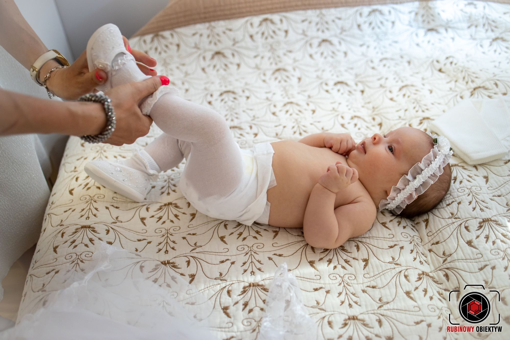 Ubrana w białe rajstopki i opaskę niemowlęca dziewczynka leży na wzorzystej kołdrze, podczas gdy osoba z neonowymi paznokciami wkłada jej białe buciki.
