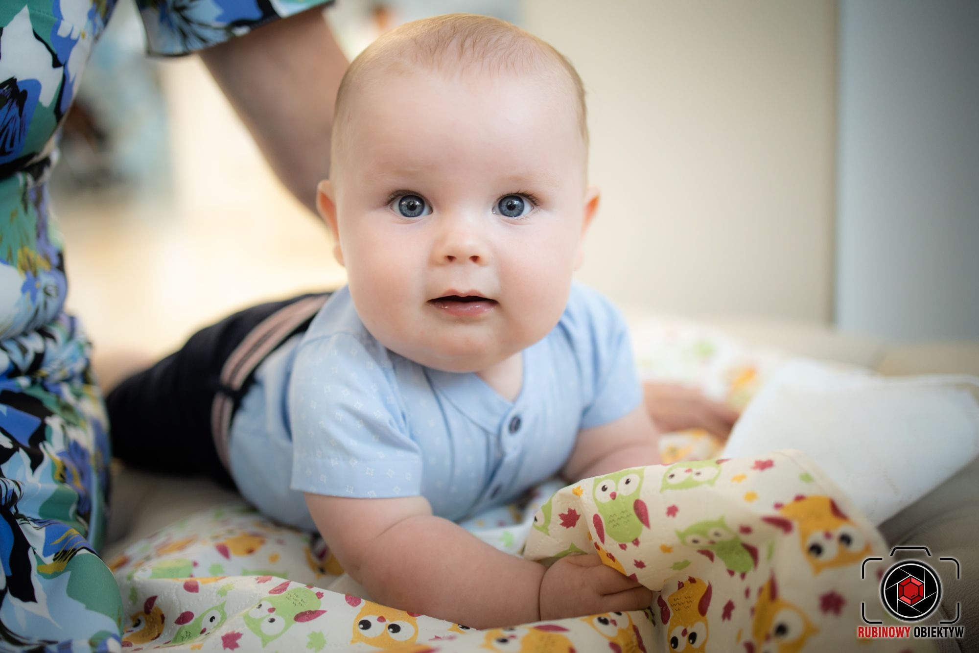 Niemowlę w błękitnym ubranku leży na brzuszku na kocyku w sowy, spoglądając w obiektyw z lekkim uśmiechem, w tle fragment kwiecistej bluzki osoby dorosłej.