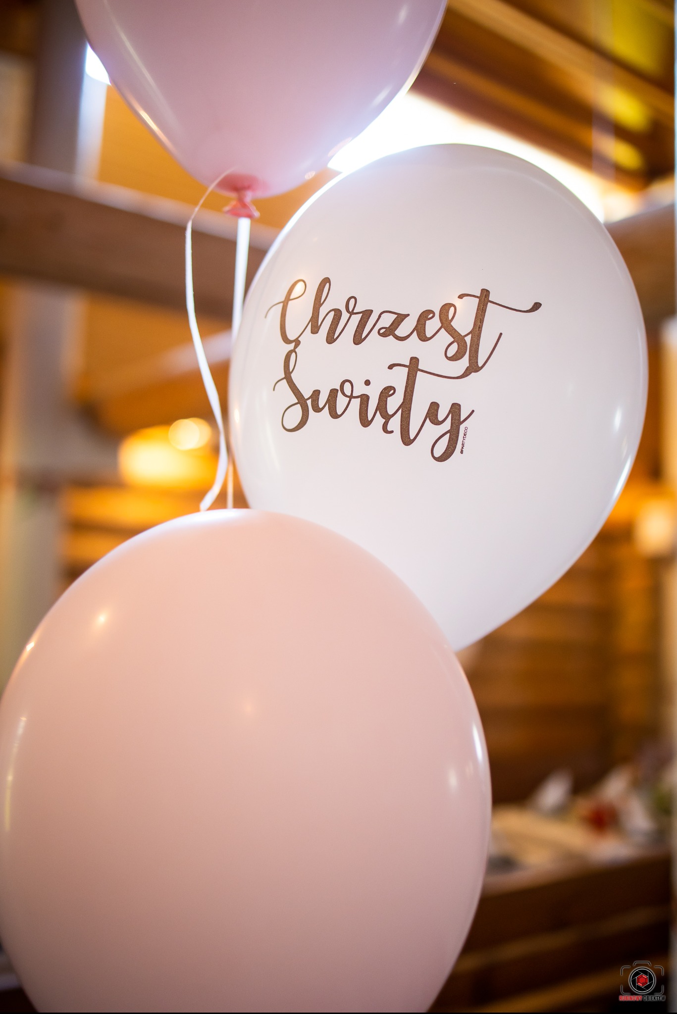 Trzy pastelowe balony: biały z napisem 'Chrzest Święty' w złotym kolorze, oraz dwa różowe, tworzące dekorację na uroczystość.