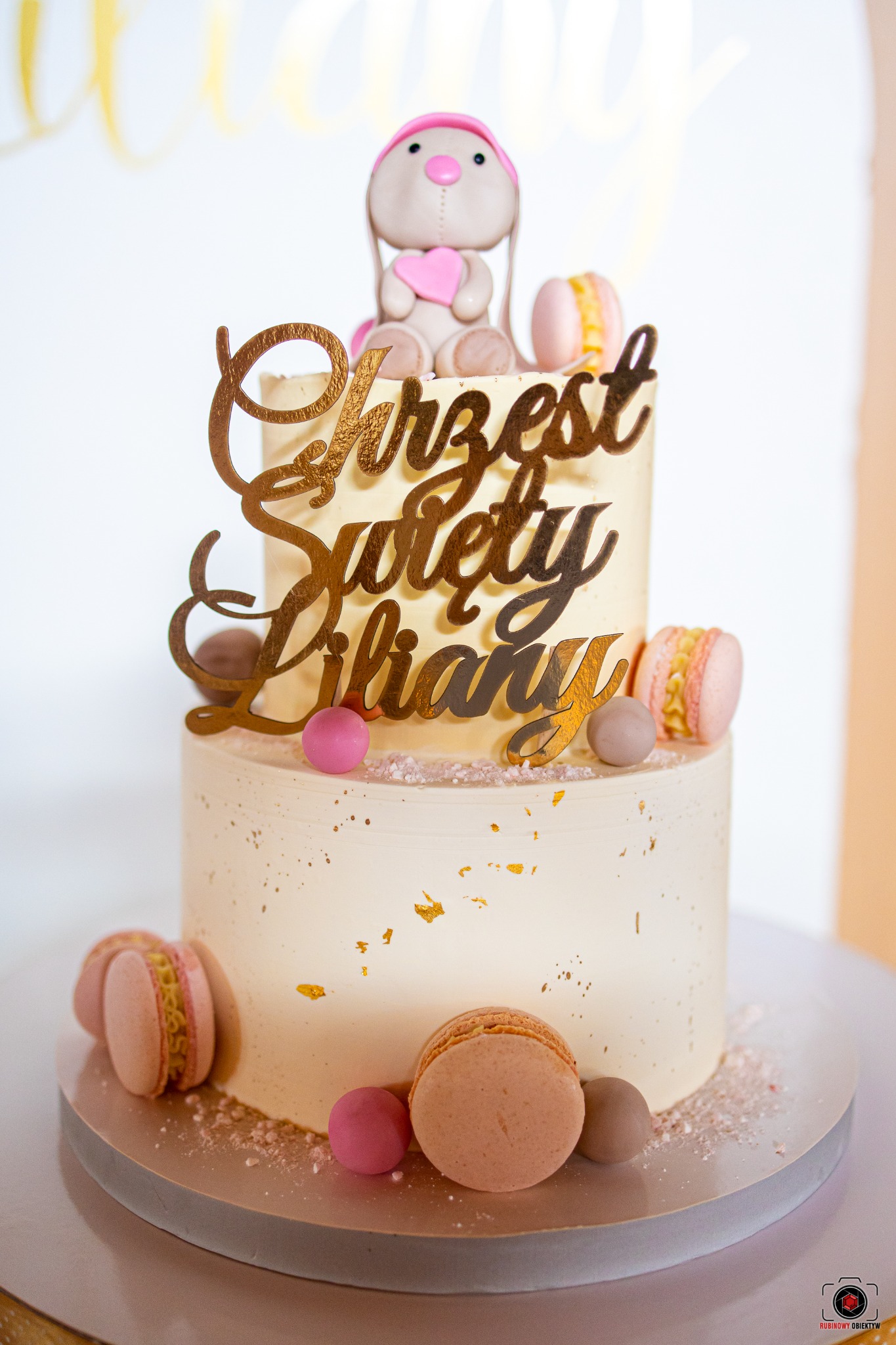 Dwupiętrowy tort w jasnych barwach, udekorowany makaronikami, złotymi płatkami i topperem z napisem 'Chrzest Święty Liliany' oraz figurką pluszowego króliczka z różowym sercem.