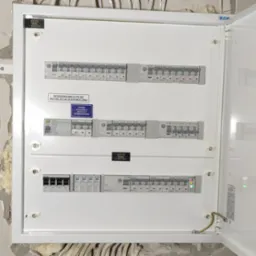 Otwarta skrzynka rozdzielcza PV AC z zainstalowanymi wyłącznikami, okablowaniem uziemiającym i naklejkami informacyjnymi, zamontowana na ścianie z widocznymi śladami prac wykończeniowych.