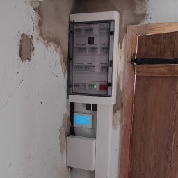 Electro Viking Sebastian Olszewski - Nowa skrzynka elektryczna z wyświetlaczem zamontowana na starej, popękanej ścianie obok drewnianych drzwi.