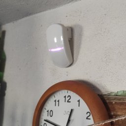 Electro Viking Sebastian Olszewski - Biały czujnik alarmowy z podświetleniem LED zamontowany na ścianie obok zegara ściennego i włącznika światła.