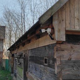 Electro Viking Sebastian Olszewski - Biała kamera monitoringu zamontowana na rogu drewnianego budynku gospodarczego, w tle drugi budynek z cegły i drewnianą konstrukcją dachu.