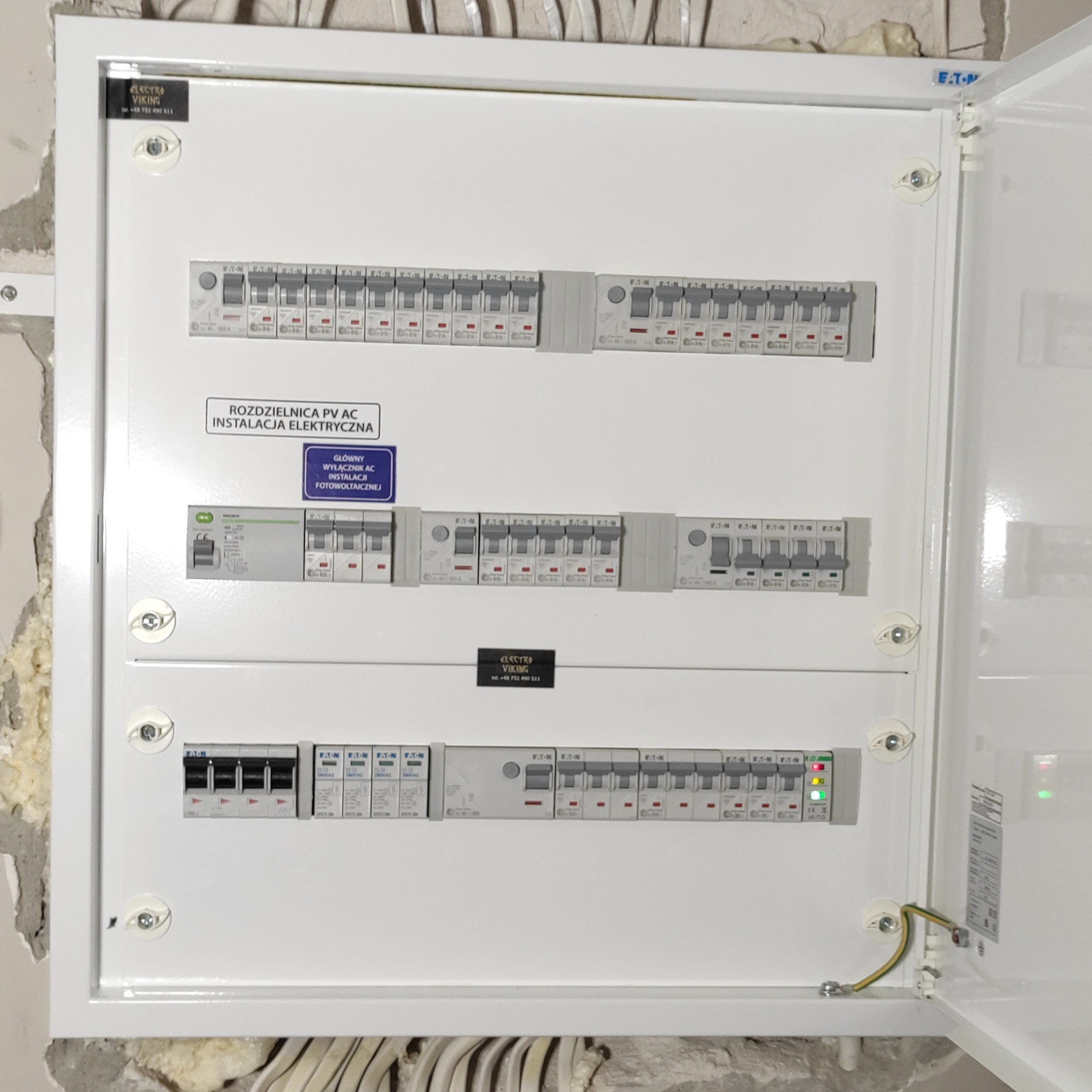 Otwarta skrzynka rozdzielcza PV AC z zainstalowanymi wyłącznikami, okablowaniem uziemiającym i naklejkami informacyjnymi, zamontowana na ścianie z widocznymi śladami prac wykończeniowych.