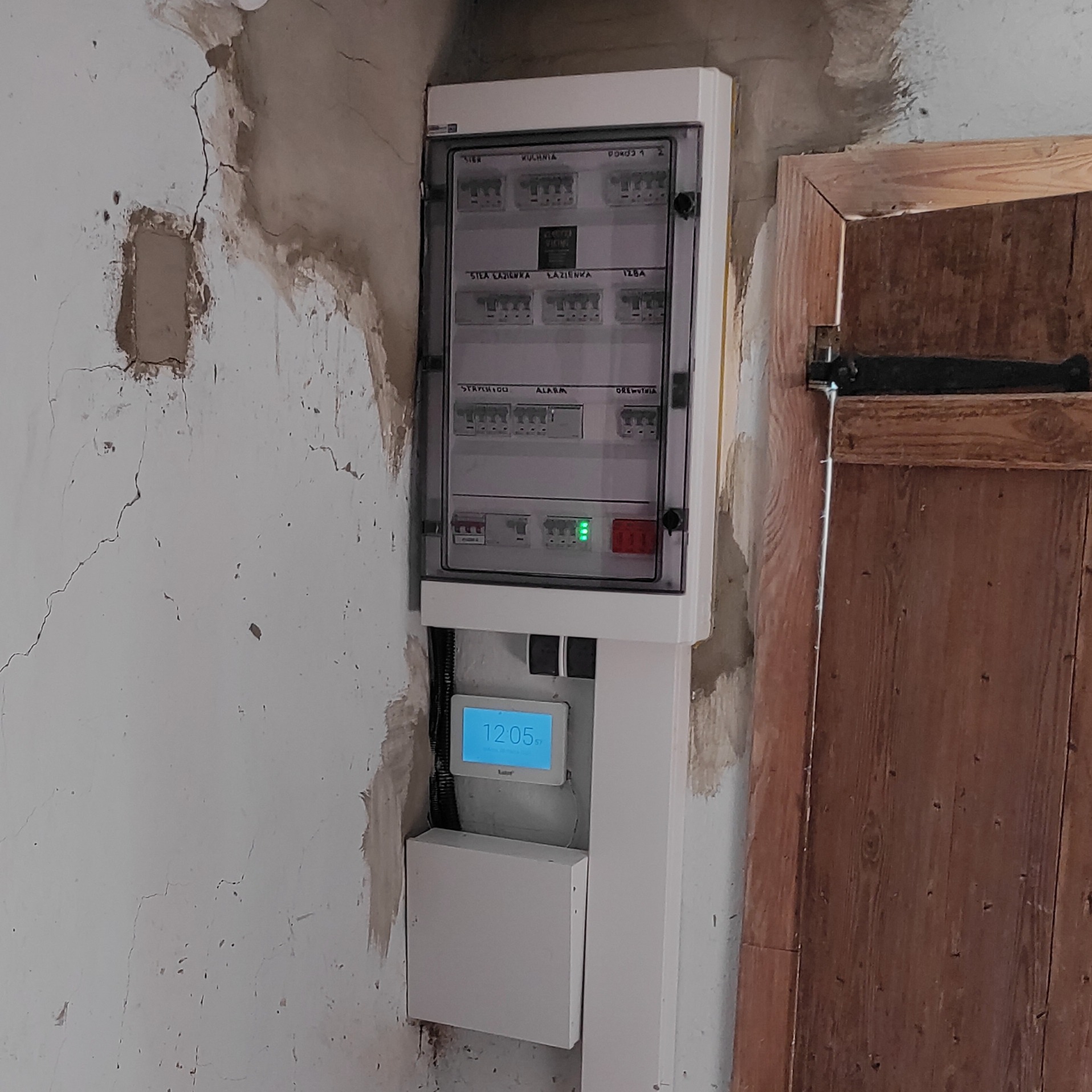 Nowa skrzynka elektryczna z wyświetlaczem zamontowana na starej, popękanej ścianie obok drewnianych drzwi.