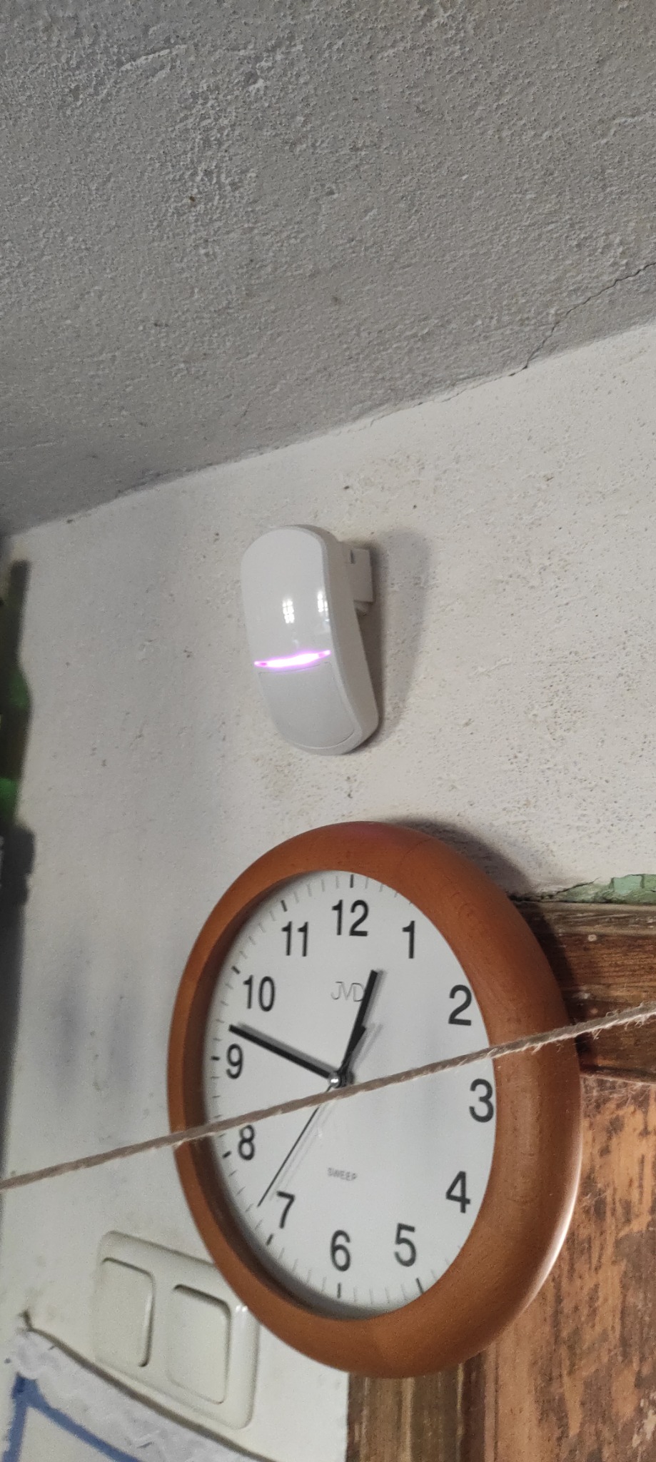 Biały czujnik alarmowy z podświetleniem LED zamontowany na ścianie obok zegara ściennego i włącznika światła.