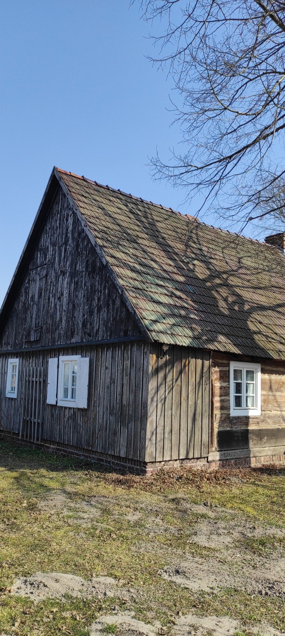 Drewniany dom z dwoma oknami z białymi ramami i brązową dachówką, widoczny z zewnątrz w słoneczny dzień. Na ścianie widać ślady upływającego czasu.