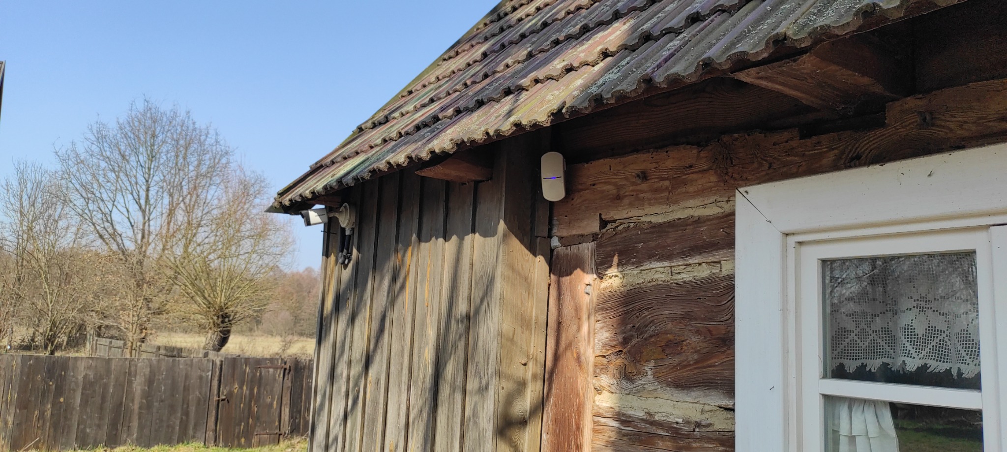 Biały czujnik alarmowy z niebieską diodą LED zamontowany na drewnianej ścianie budynku z oknem, widoczne również elementy systemu alarmowego.