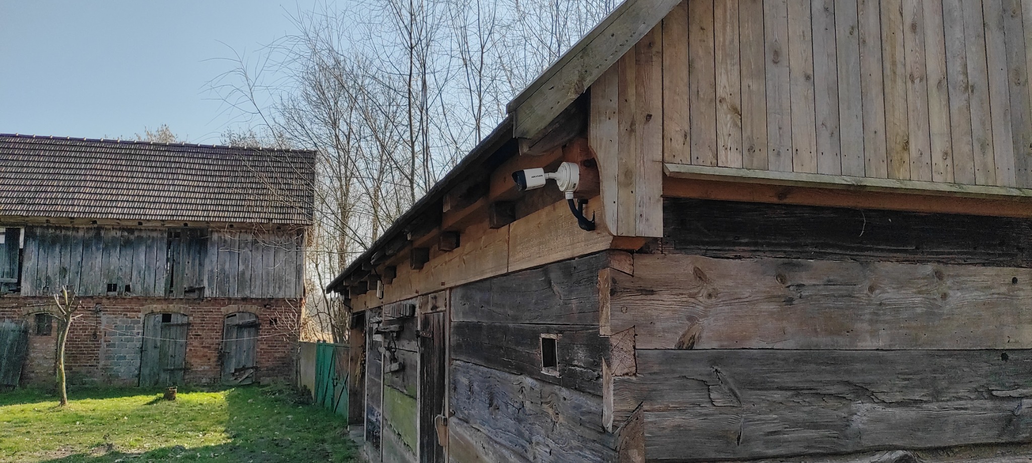 Biała kamera monitoringu zamontowana na rogu drewnianego budynku gospodarczego, w tle drugi budynek z cegły i drewnianą konstrukcją dachu.