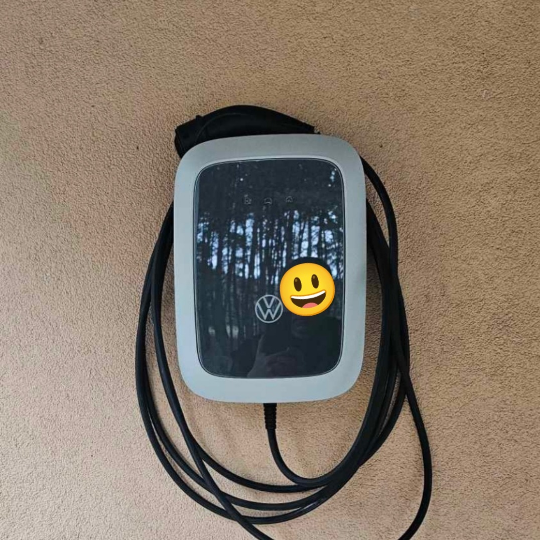 Szara stacja ładowania samochodów elektrycznych Volkswagen zamontowana na teksturowanej ścianie, z widocznym kablem i odbiciem otoczenia na wyświetlaczu.