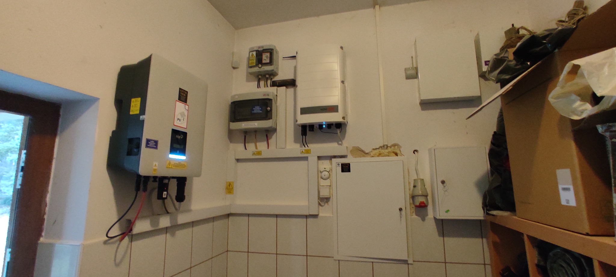 Wnętrze pomieszczenia z zamontowanymi elementami instalacji fotowoltaicznej: inwertery, zabezpieczenia, okablowanie, widoczne płytki ceramiczne na ścianach.