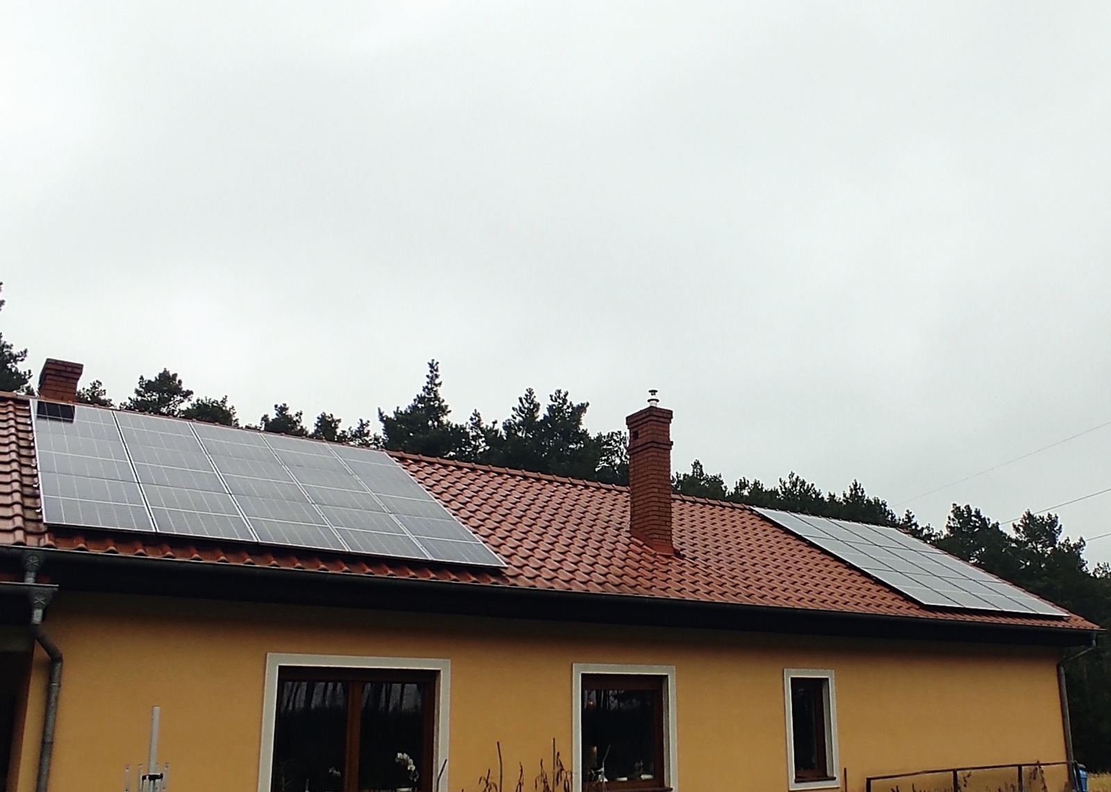 Panele słoneczne zamontowane na dachu domu z czerwoną dachówką, widoczne dwa kominy i drzewa w tle pod pochmurnym niebem.