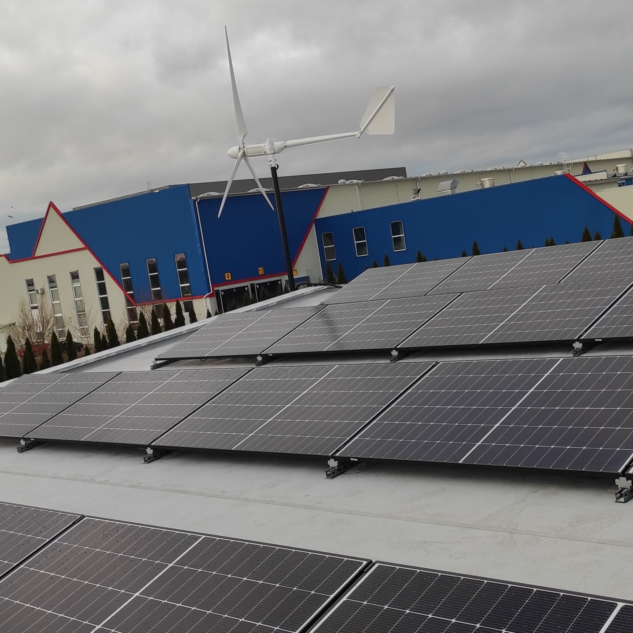 Instalacja fotowoltaiczna na dachu budynku przemysłowego z turbiną wiatrową w tle, widoczne panele słoneczne zamontowane na szarym dachu, pochmurne niebo.