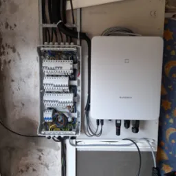 Otwarta skrzynka rozdzielcza z widocznymi bezpiecznikami i okablowaniem, obok zamontowany biały inwerter marki Sungrow, kable w peszlach prowadzone wzdłuż ściany.