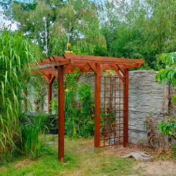 Drewniana pergola z kratką w ogrodzie, obok oczko wodne i ozdobne trawy. Konstrukcja z ciemnego drewna na tle kamiennego murku i bujnej zieleni.