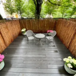 Umeblowany balkon z podłogą z desek kompozytowych, otoczony bambusową matą, z kwiatami w donicach i metalowym zestawem mebli ogrodowych.