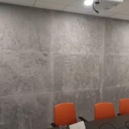 Ściana w biurze wykończona szarą, dekoracyjną gładzią imitującą kamień, podzielona na kwadratowe panele, z widocznym projektorem na suficie i pomarańczowymi krzesłami konferencyjnymi.