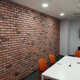 Sala konferencyjna z dekoracyjną ścianą z czerwonej cegły, białym stołem i pomarańczowymi krzesłami, oświetlona lampami sufitowymi i z projektorem na suficie.