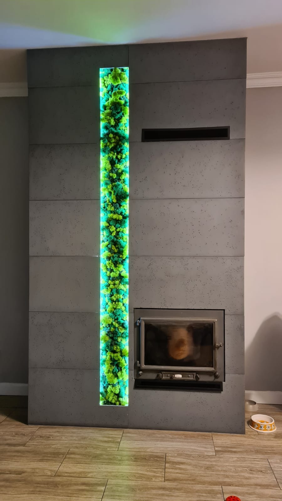 Szary betonowy kominek z wbudowanym oświetleniem LED i panelem z mchu stabilizowanego, odbicie twarzy w szybie kominka.
