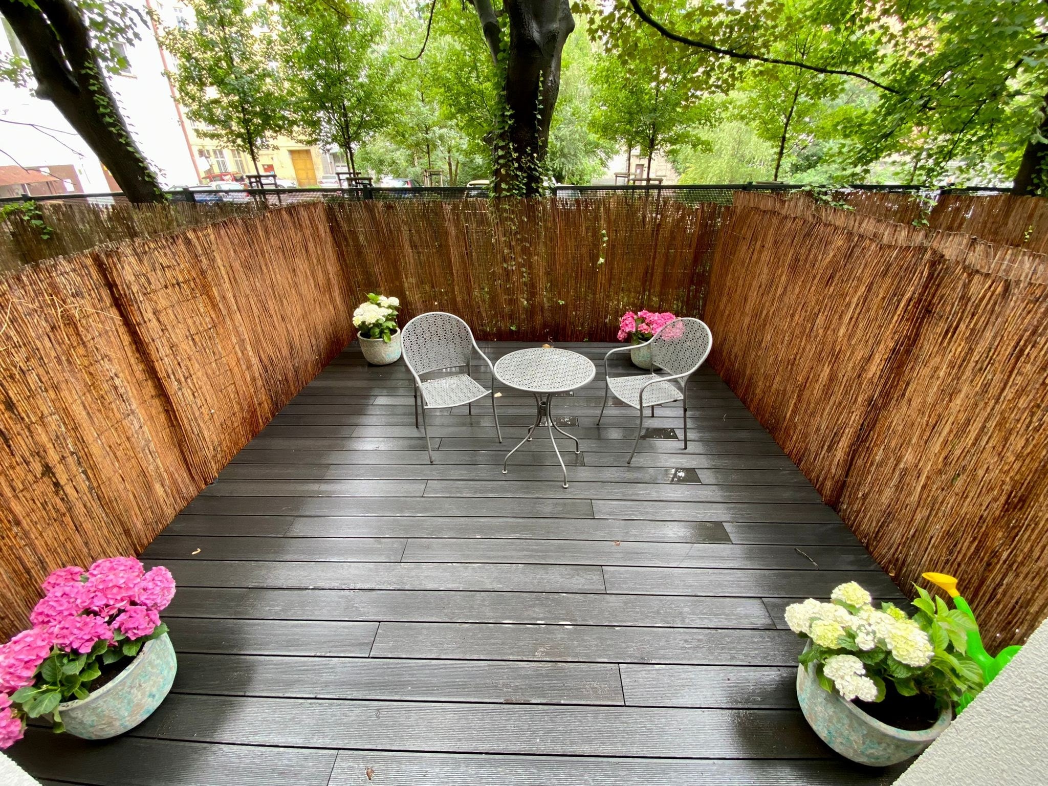 Umeblowany balkon z podłogą z desek kompozytowych, otoczony bambusową matą, z kwiatami w donicach i metalowym zestawem mebli ogrodowych.