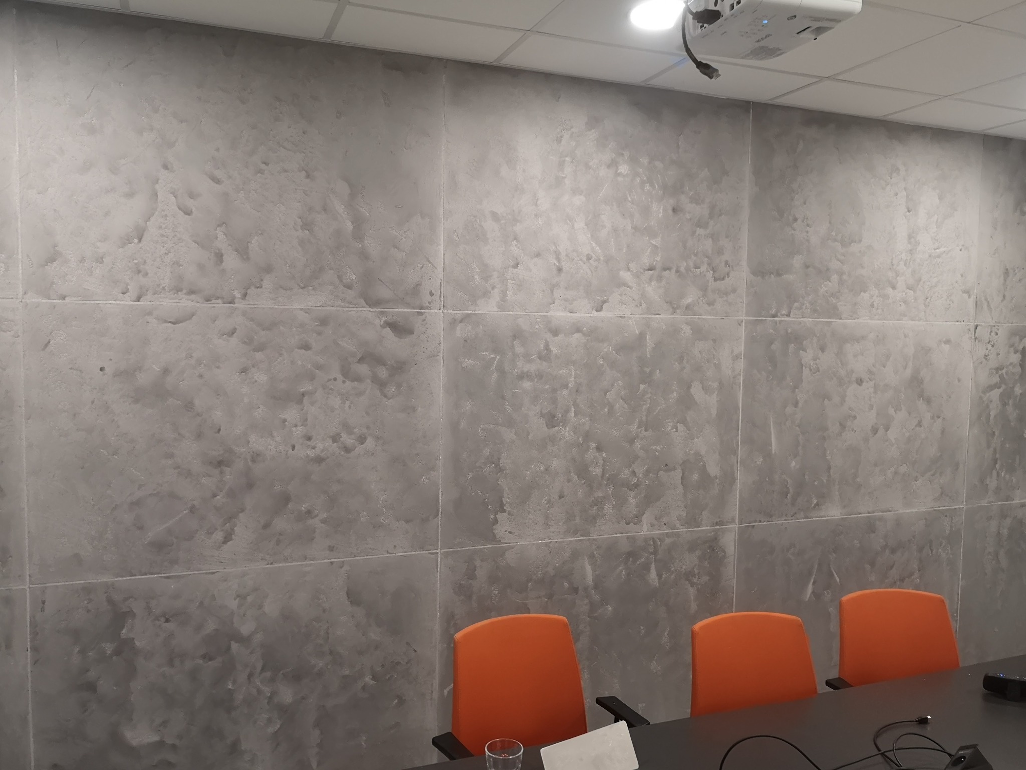 Ściana w biurze wykończona szarą, dekoracyjną gładzią imitującą kamień, podzielona na kwadratowe panele, z widocznym projektorem na suficie i pomarańczowymi krzesłami konferencyjnymi.