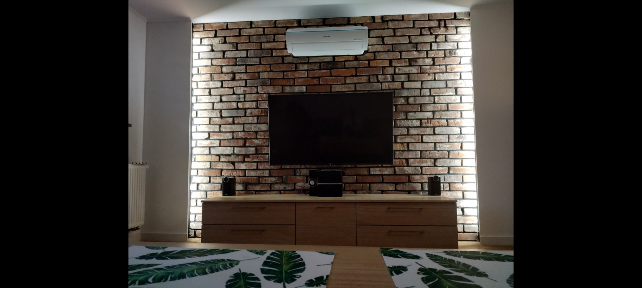 Telewizor zawieszony na ścianie z cegły klinkierowej, podświetlonej pionowymi pasami LED, nad drewnianą komodą z szufladami i sprzętem audio.