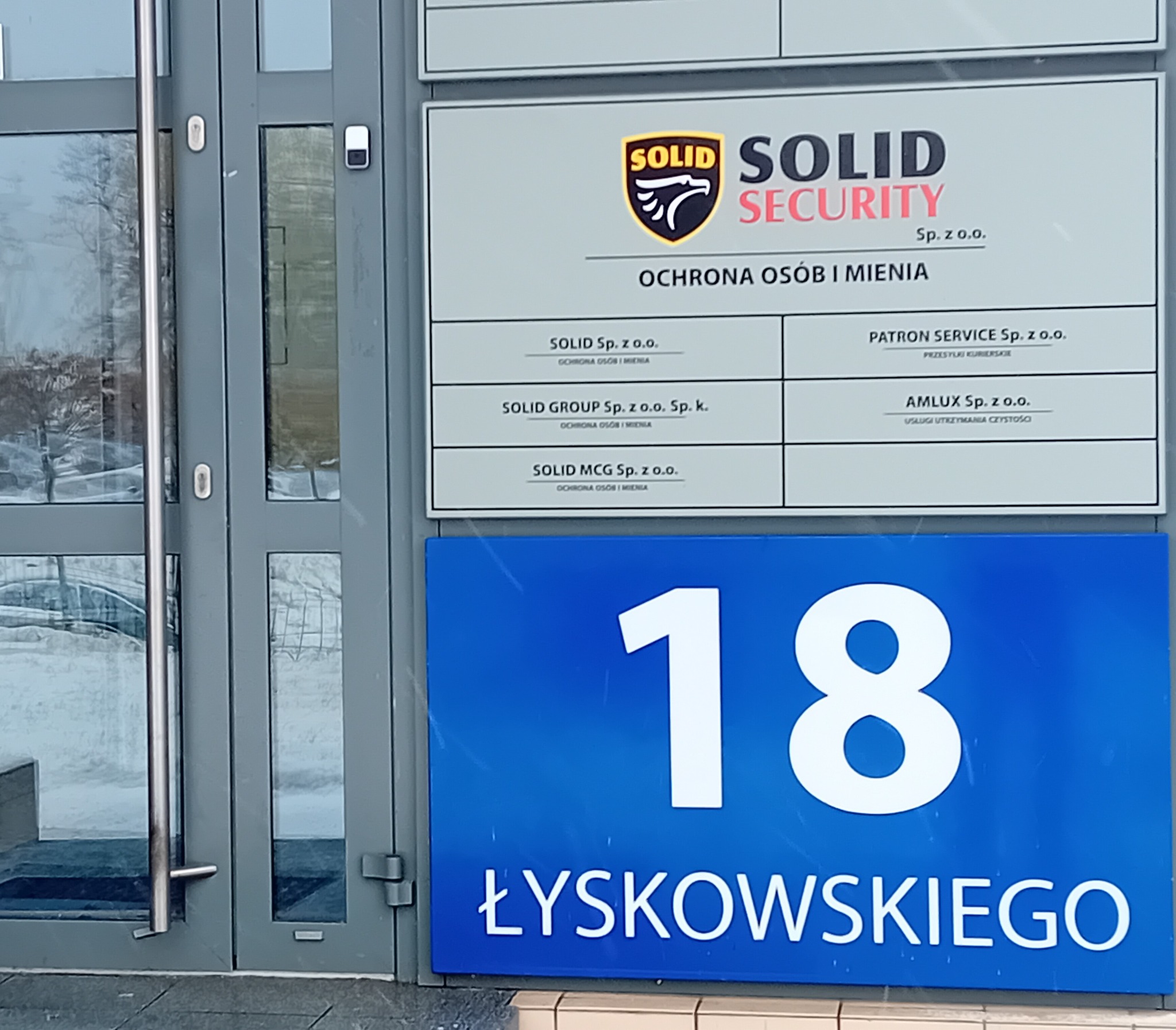 Tablica informacyjna z nazwą i logo firmy Solid Security Sp. z o.o. oraz listą innych firm powiązanych, umieszczona nad niebieską tabliczką z numerem budynku 18 przy ulicy Łyskowskiego, obok...