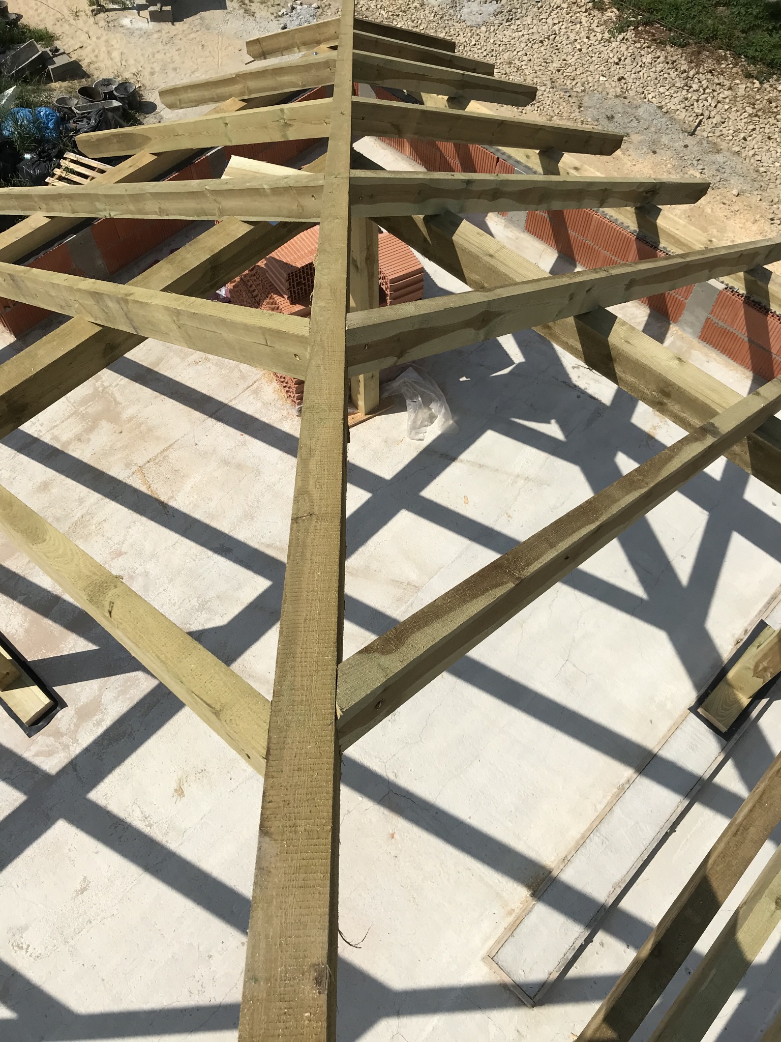 Drewniana konstrukcja więźby dachowej z widocznymi elementami muru z czerwonej cegły i zapasem dachówek w tle, oświetlona jasnym światłem słonecznym tworzącym ostre cienie.