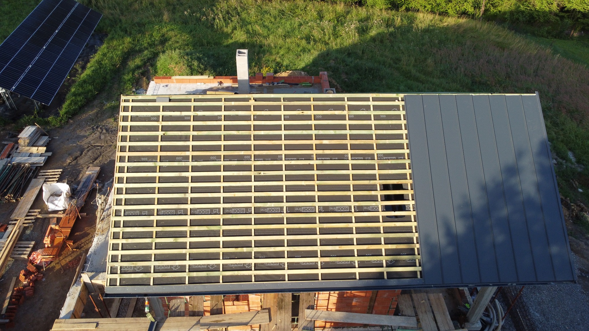 Widok z góry na konstrukcję dachu w trakcie budowy, z widoczną więźbą, membraną dachową z nadrukiem 'ROOFline' oraz częściowo ułożonym grafitowym pokryciem z blachy na rąbek stojący, w tle komin...