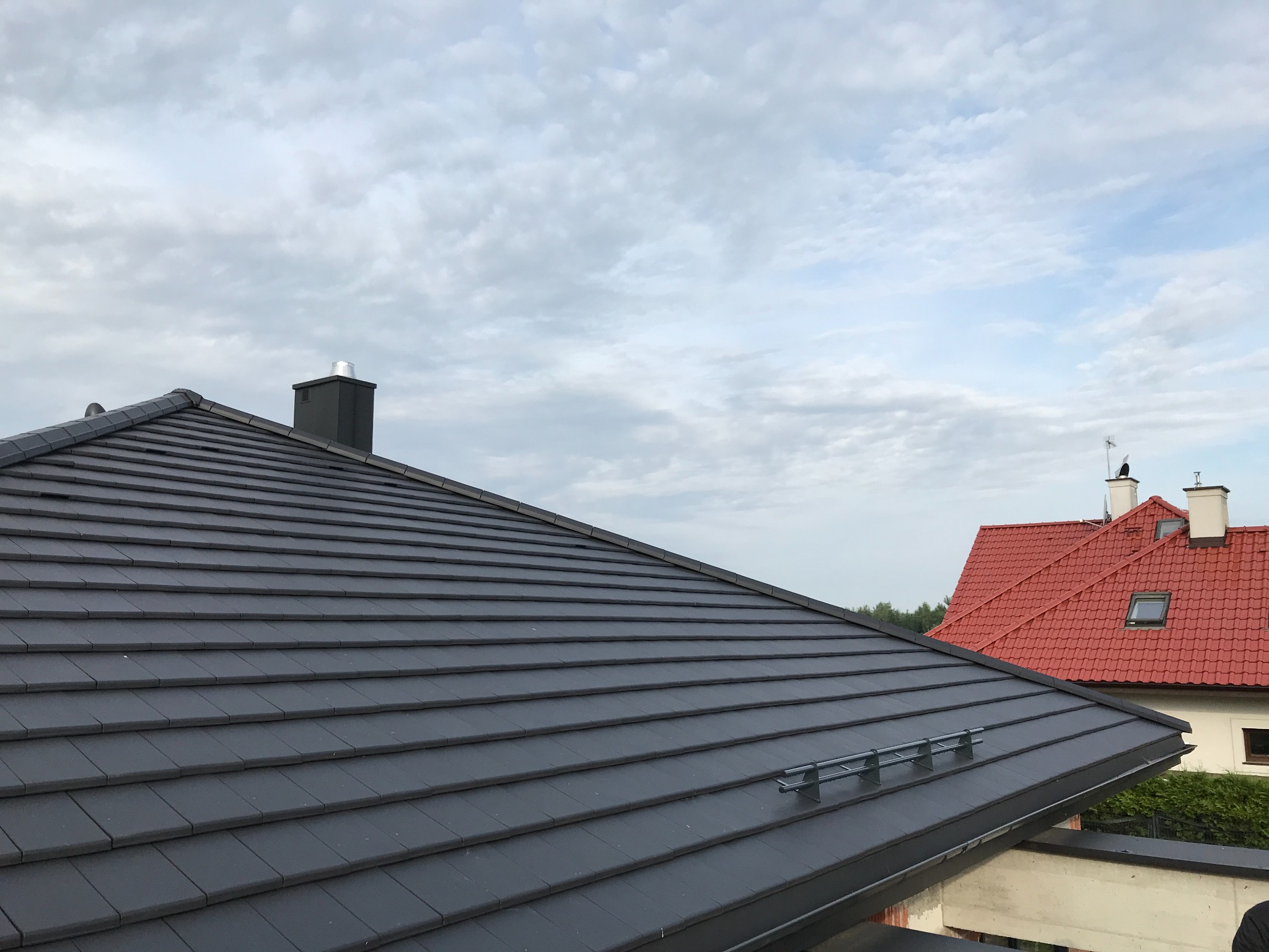 Ciemnoszary dach z dachówki Creaton Simpla, widoczne zabezpieczenia przeciwśniegowe, w tle dach sąsiedniego budynku z czerwonej dachówki i komin.