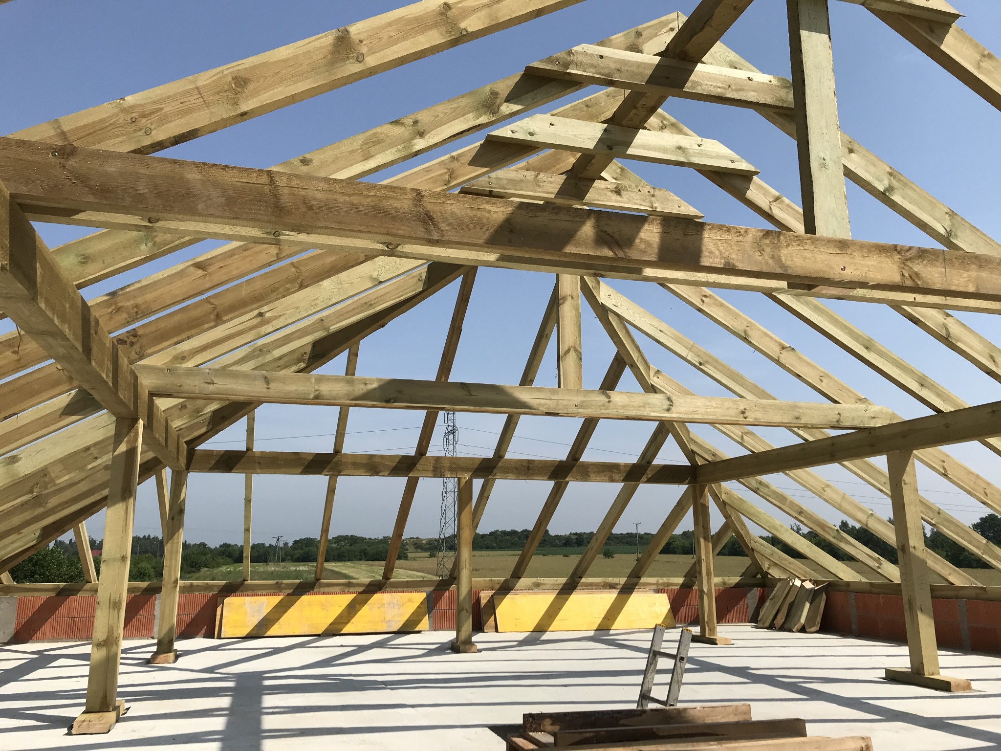 Wznoszenie drewnianej więźby dachowej na murowanym budynku, widok na konstrukcję z belek pod kątem, z elementami rusztowania i krajobrazem w tle.