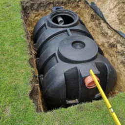 Czarny zbiornik retencyjny z rurą w wykopie ziemnym na trawniku, z poziomnicą w pobliżu króćca.