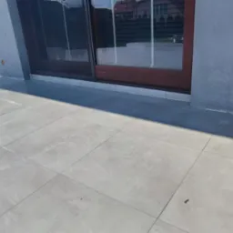 Świeżo położone, jasnoszare płytki na tarasie zewnętrznym, częściowo mokre po myciu, z widocznymi kolumnami i oknem przesuwnym w tle.