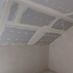 RBTS Usługi Ogólnobudowlane Robert Szczepańczyk - Montaż płyt gipsowo-kartonowych na skośnym suficie poddasza, widoczne łączenia płyt zaszpachlowane, okno z widokiem na dachy sąsiednich budynków.