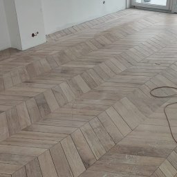 RBTS Usługi Ogólnobudowlane Robert Szczepańczyk - Ułożona podłoga z płytek imitujących drewno w jodełkę, widoczny kabel i wiadro w tle, białe ściany z dekoracyjną listwą.