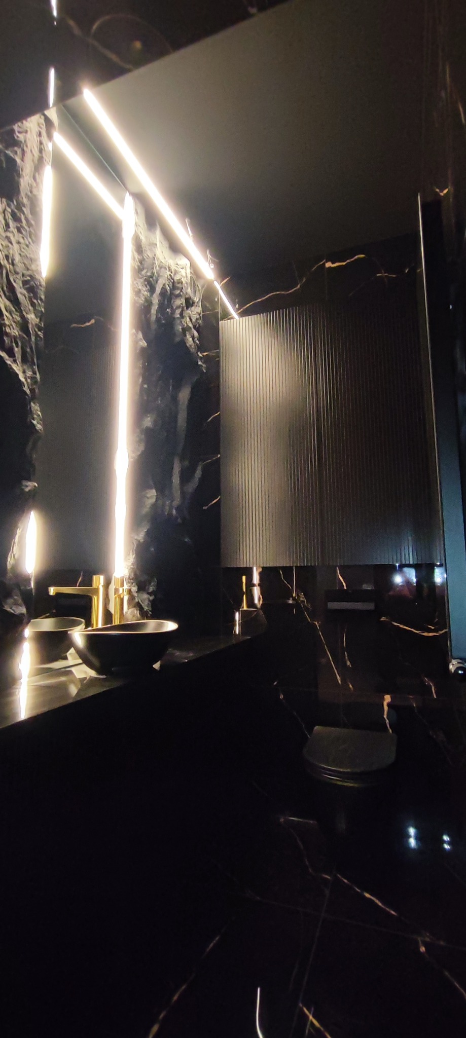 Nowoczesna łazienka z czarnym marmurem, podświetlanymi ścianami o fakturze skały i złotą armaturą, ukazująca eleganckie wykończenie.